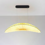 A Clérachic pendant with a black oval ceiling plate and a pale pleated shade that glows yellow in the middle, against a plain wall.