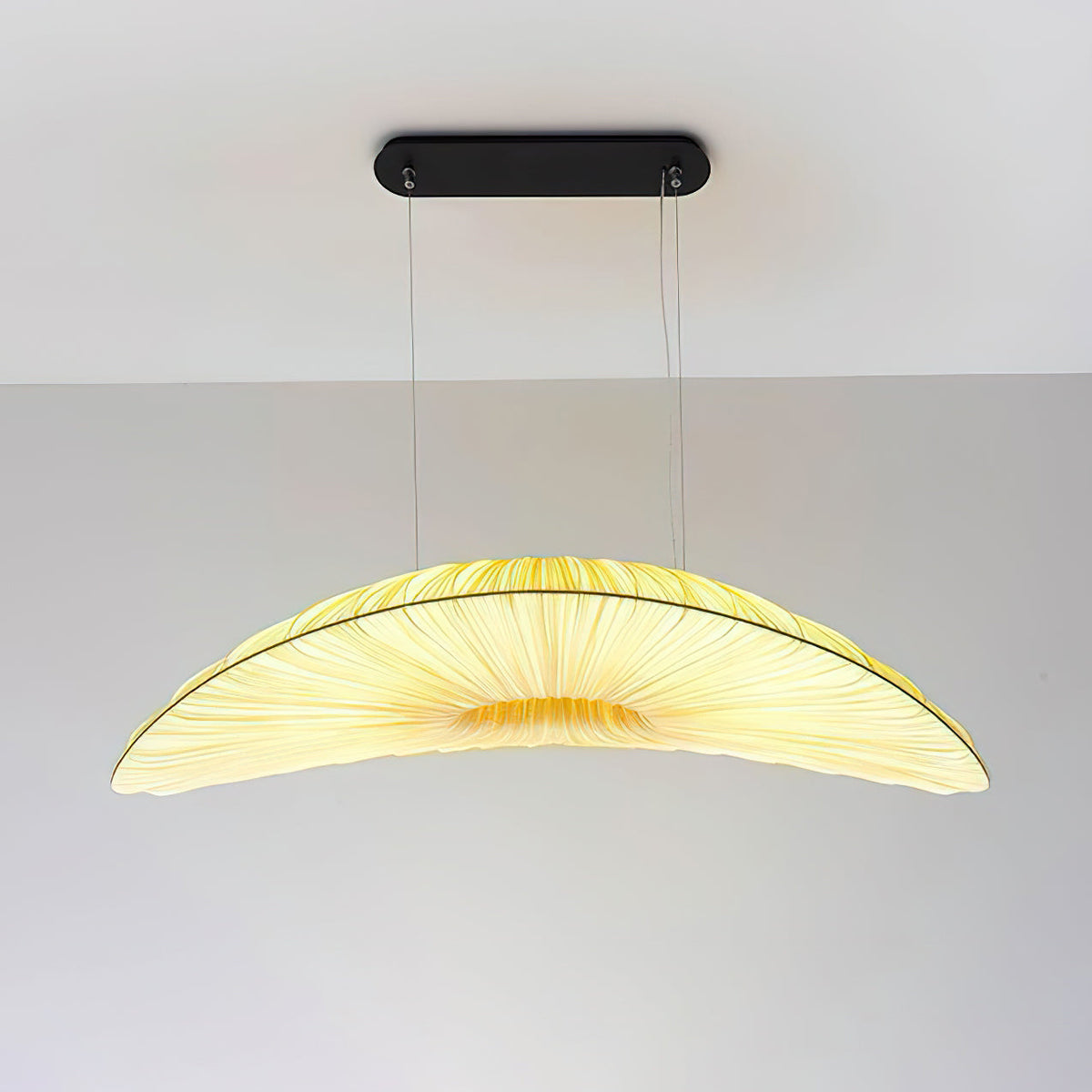 A Clérachic pendant with a black oval ceiling plate and a pale pleated shade that glows yellow in the middle, against a plain wall.