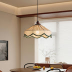 Lit Clérachic pleated beige pendant above a round dark table with a bowl of lemons, beside a window with white blinds.