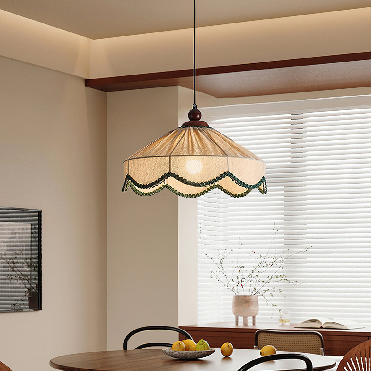 Lit Clérachic pleated beige pendant above a round dark table with a bowl of lemons, beside a window with white blinds.