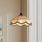 Close view of a lit Clérachic pendant, its pleated beige shade edged in green scalloped trim, beside cream curtains.