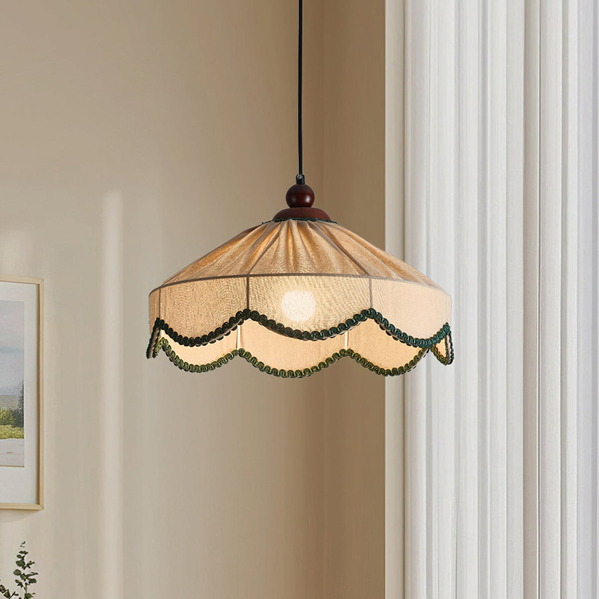 Close view of a lit Clérachic pendant, its pleated beige shade edged in green scalloped trim, beside cream curtains.