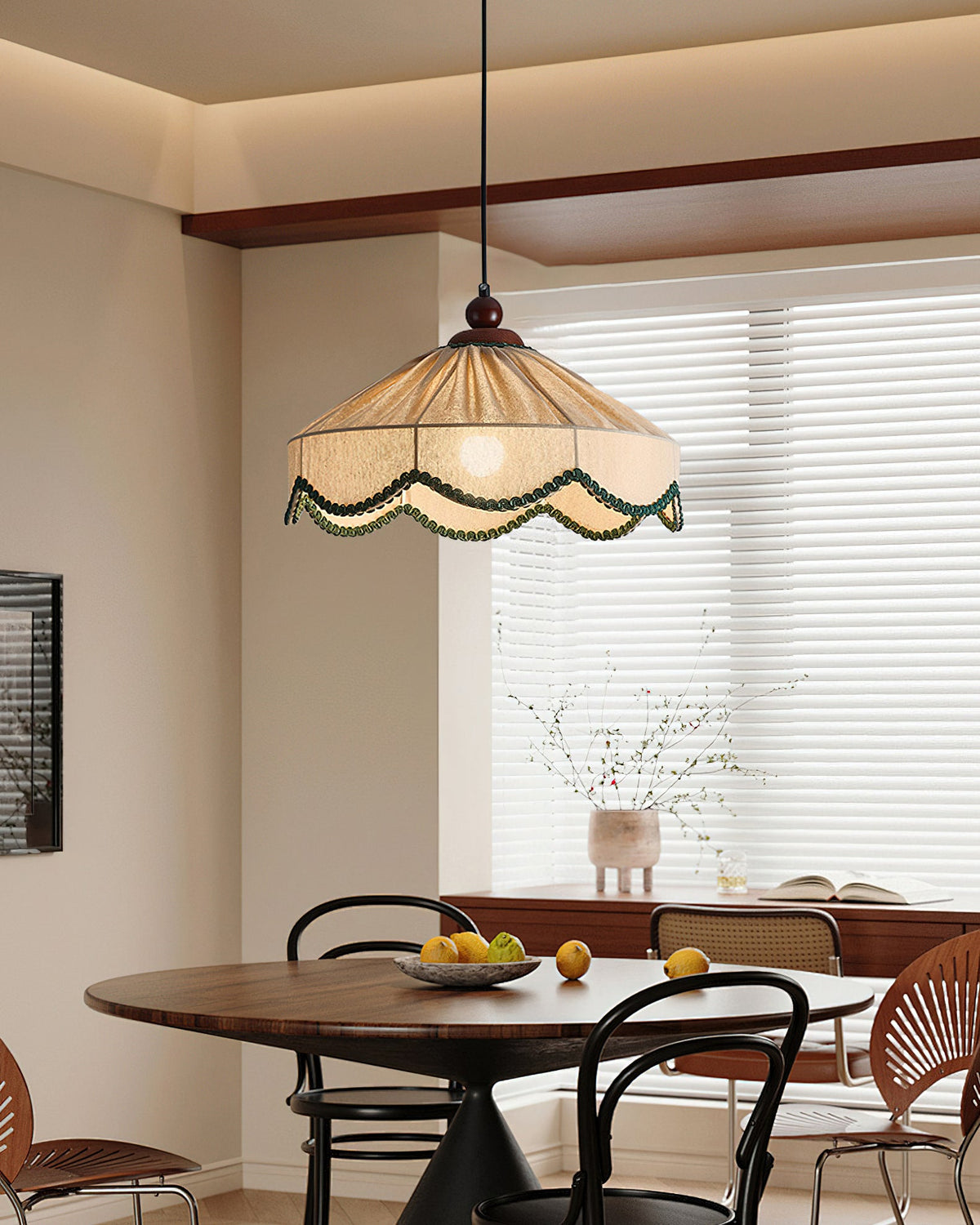 Clérachic pleated beige pendant hung over a round dark table with bentwood chairs, in front of a window with white blinds.