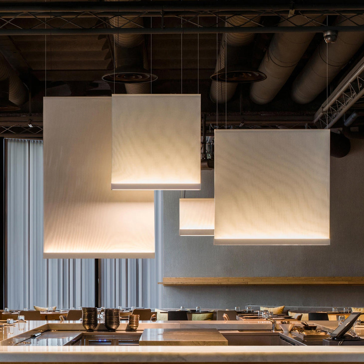 Several Clérachic fabric panel pendants of different sizes lit at staggered heights in an industrial space with exposed ducts, above an open kitchen counter.