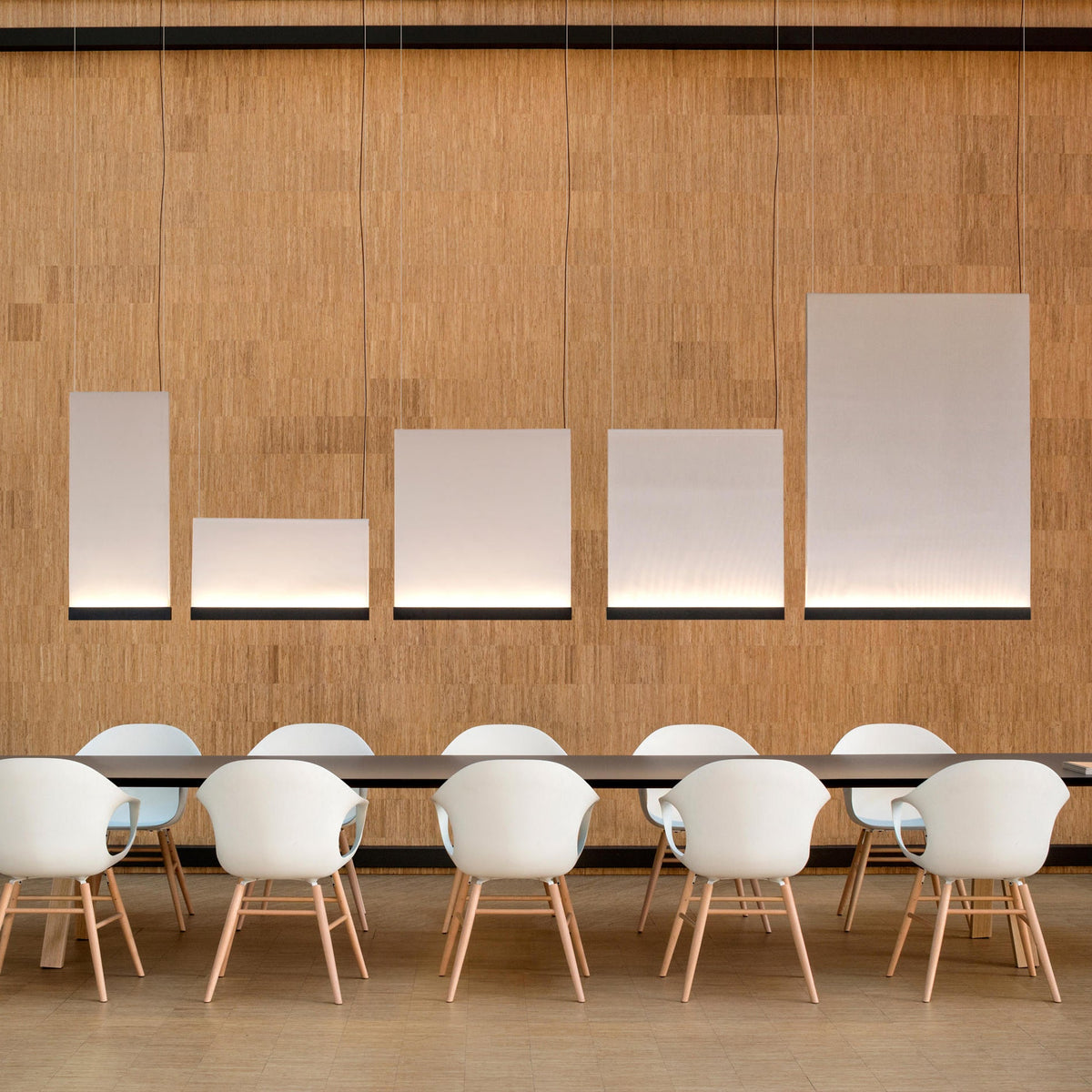 Five Clérachic fabric panel pendants of different shapes hung in a line above a long table with white shell chairs, against a cork-textured wall.