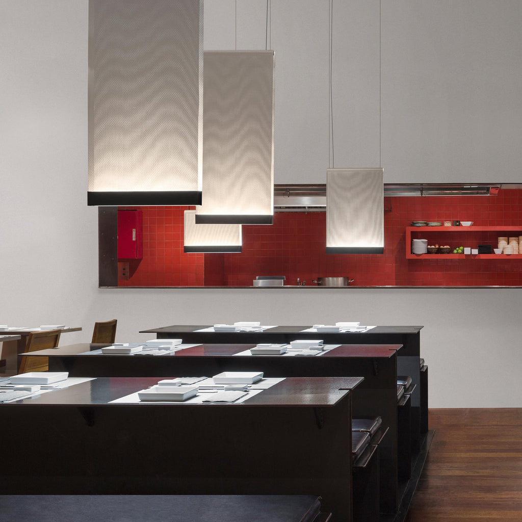 Three tall Clérachic fabric pendants lit above black tables, in front of a red tiled kitchen wall.