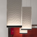 Close, low view of two tall Clérachic fabric pendants with black lower bars, glowing softly in front of red wall tiles.