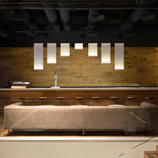 Seven Clérachic fabric panel pendants of varied lengths hung in a row above a taupe sofa and a wood-clad wall, with a bar counter behind.