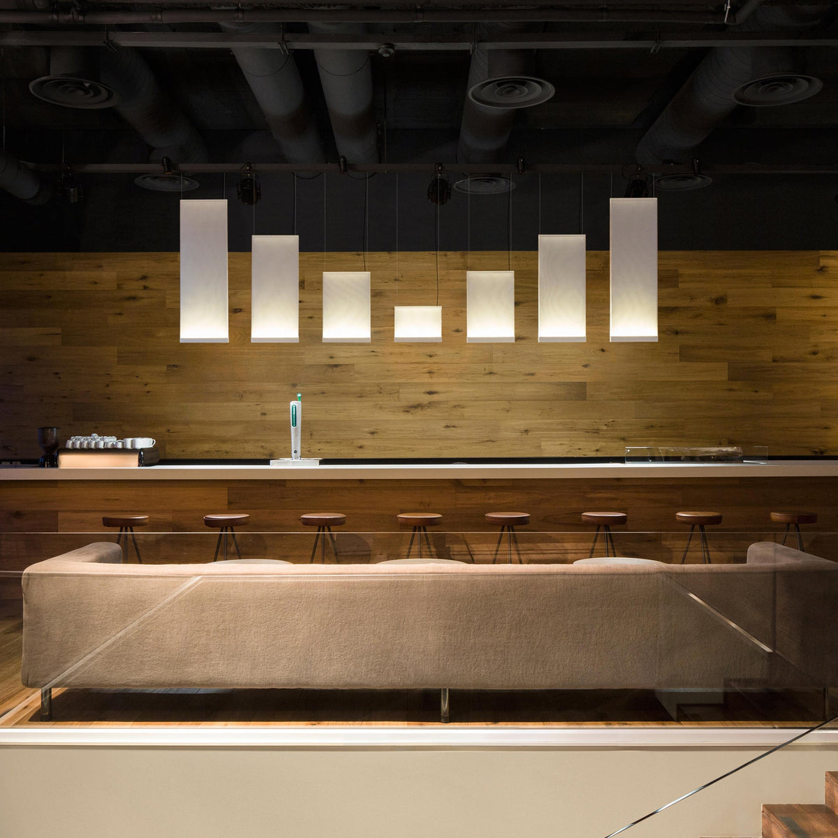 A row of Clérachic fabric panel pendants of different lengths lit above a long taupe sofa, against a warm oak-clad wall.
