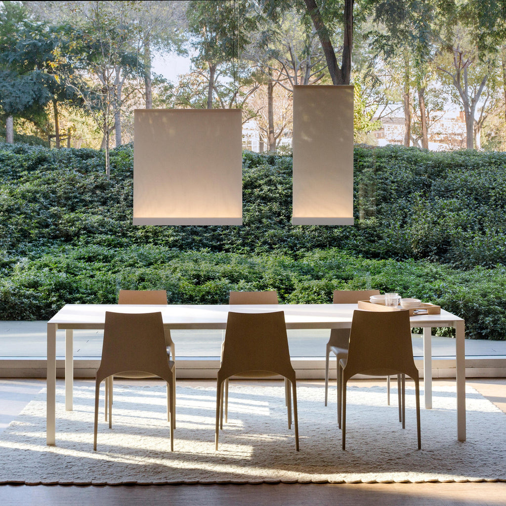 Two Clérachic fabric pendants lit in daylight over an outdoor dining table with tan chairs, with a hedge and trees behind.