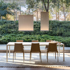 Two Clérachic fabric pendants lit in daylight over an outdoor dining table with tan chairs, with a hedge and trees behind.
