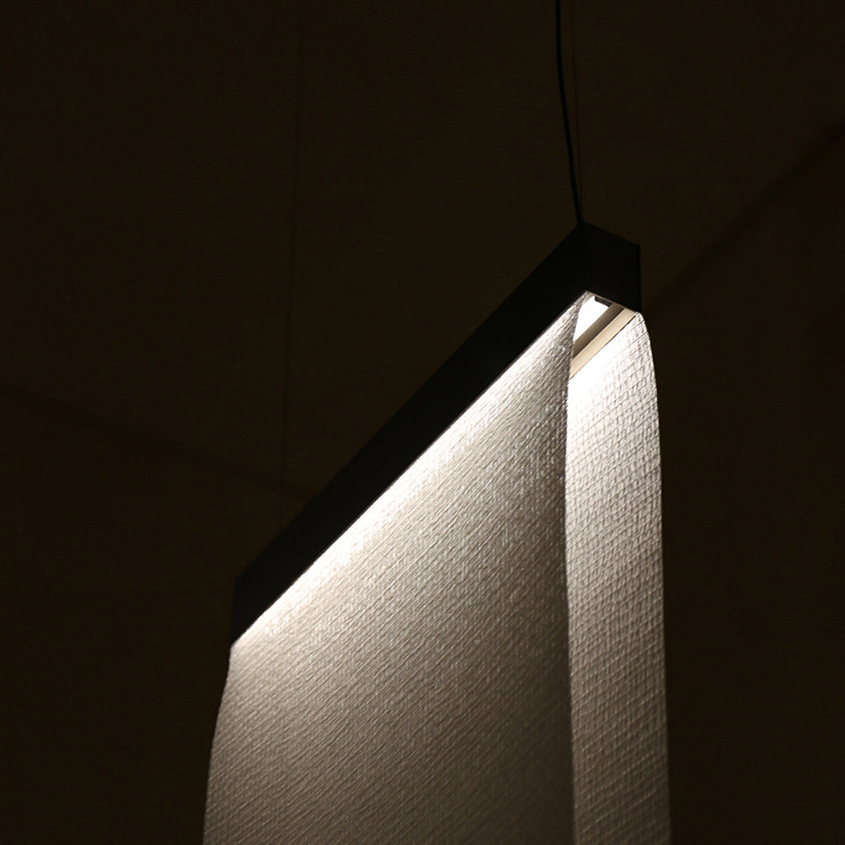 Close view of a Clérachic fabric panel curving down from a dark top bar, with a woven texture lit from the edge in a dark room.