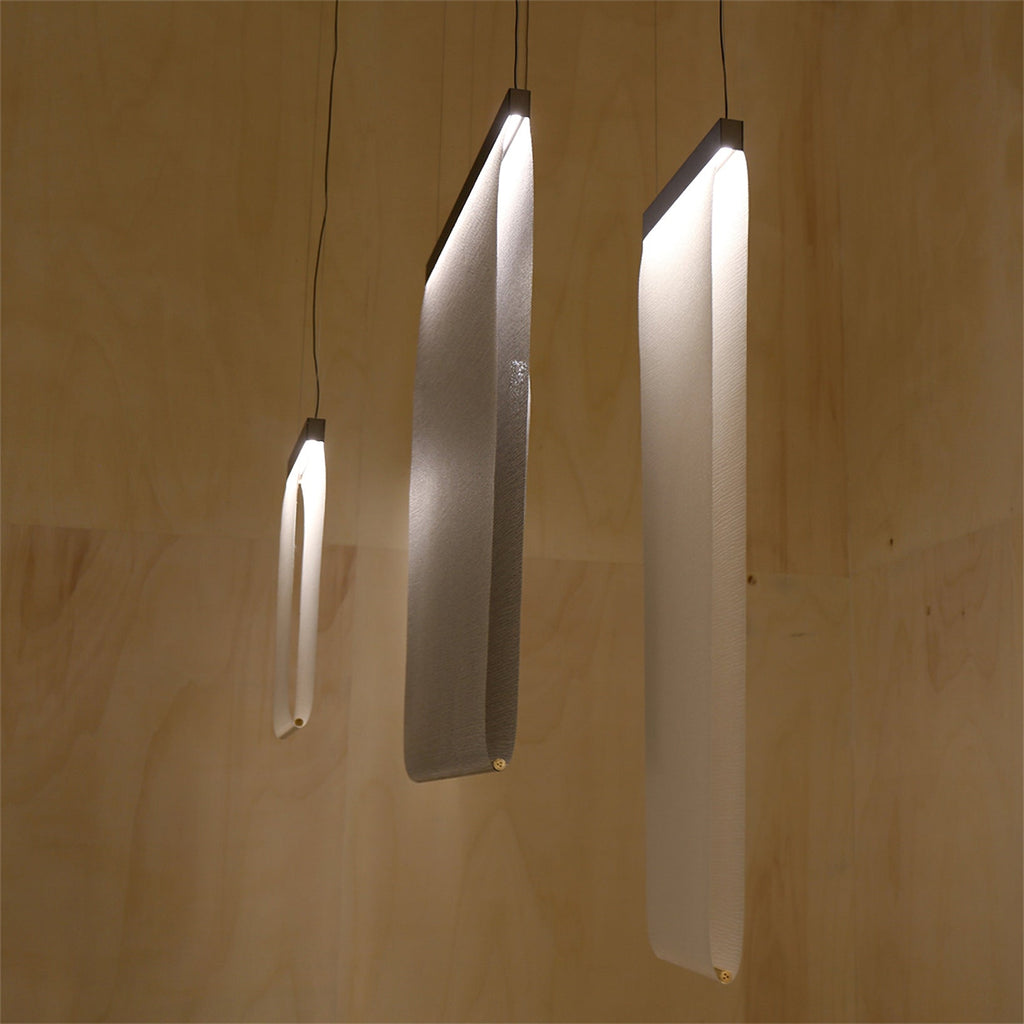 Three Clérachic fabric strips lit and curved at different angles, hung on single cords against a pale plywood-lined wall.
