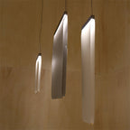 Three Clérachic fabric strips lit and curved at different angles, hung on single cords against a pale plywood-lined wall.