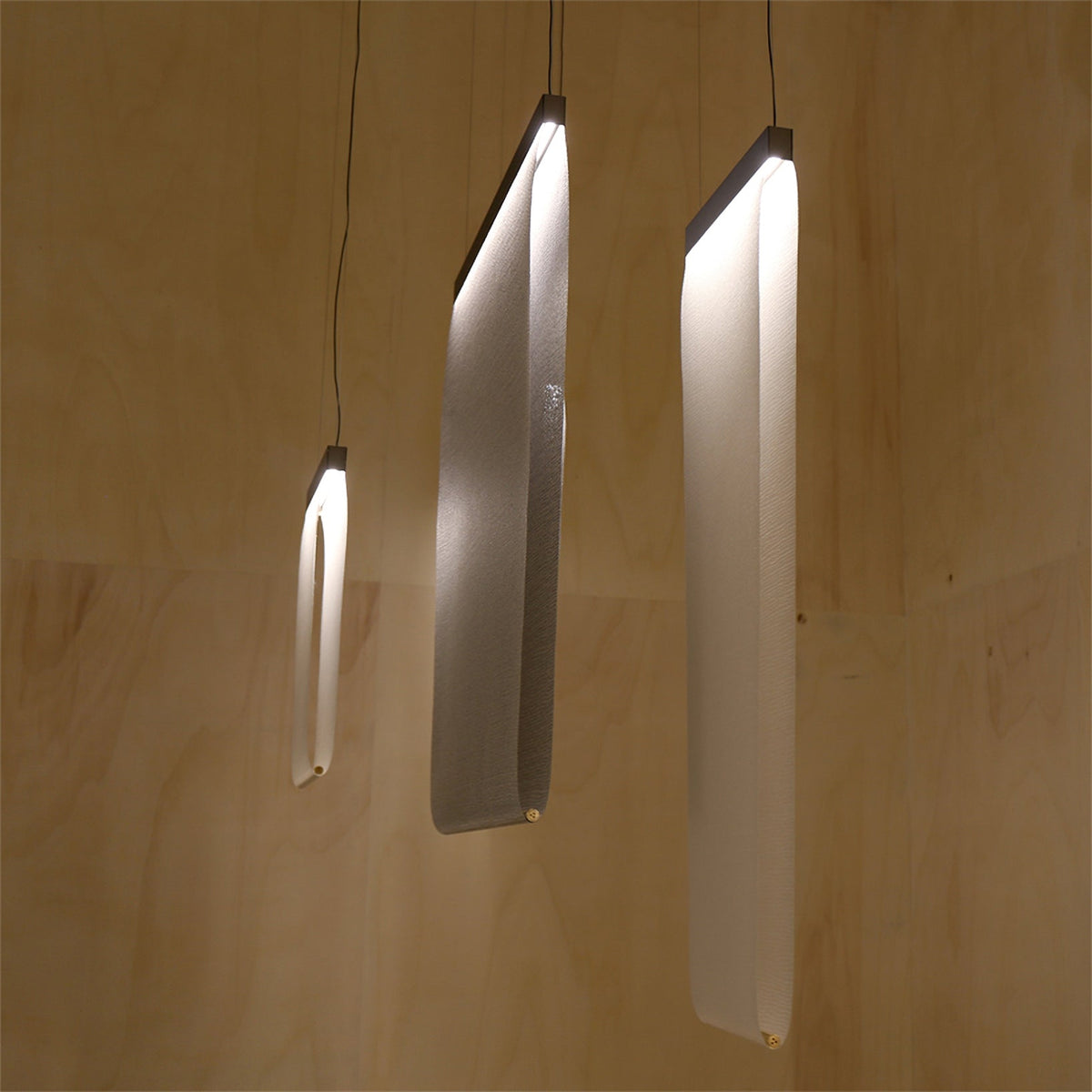Three Clérachic fabric strips lit and curved at different angles, hung on single cords against a pale plywood-lined wall.