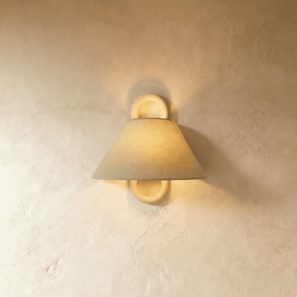 Clérachic wall light with a sage-toned fabric shade and solid wood ring mount, glowing warmly against a plaster bedroom wall.