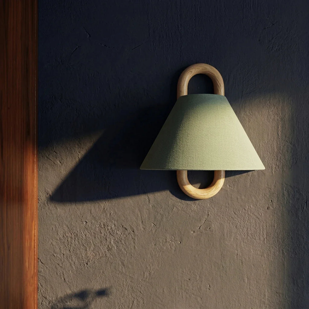 A Clérachic sage green wall light glows beside a mirror in a dim, dark-walled bathroom at dusk.