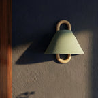 A Clérachic sage green wall light glows beside a mirror in a dim, dark-walled bathroom at dusk.