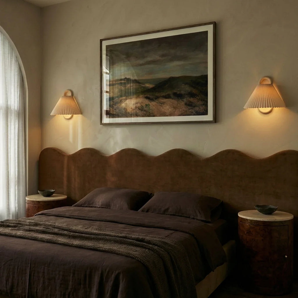 Two Clérachic pleated cream wall lights flank a landscape painting, glowing softly above a curved bedroom headboard.