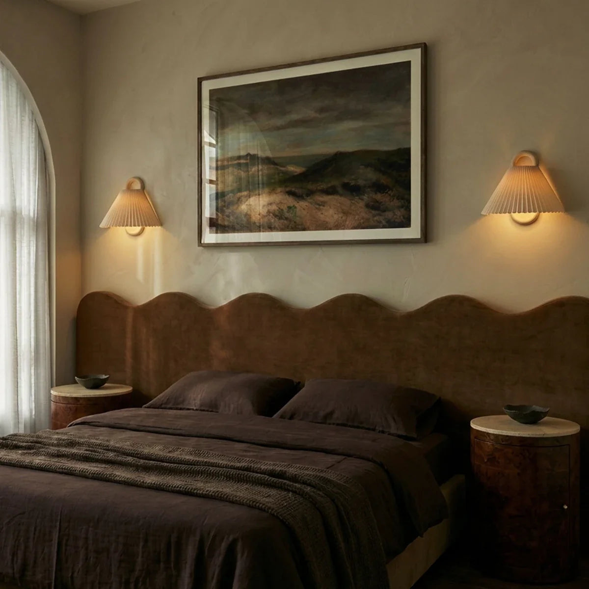 Two Clérachic pleated cream wall lights flank a landscape painting, glowing softly above a curved bedroom headboard.