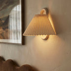 Close-up of a Clérachic pleated cream fabric shade and wood ring bracket, glowing beside a framed painting on the wall.
