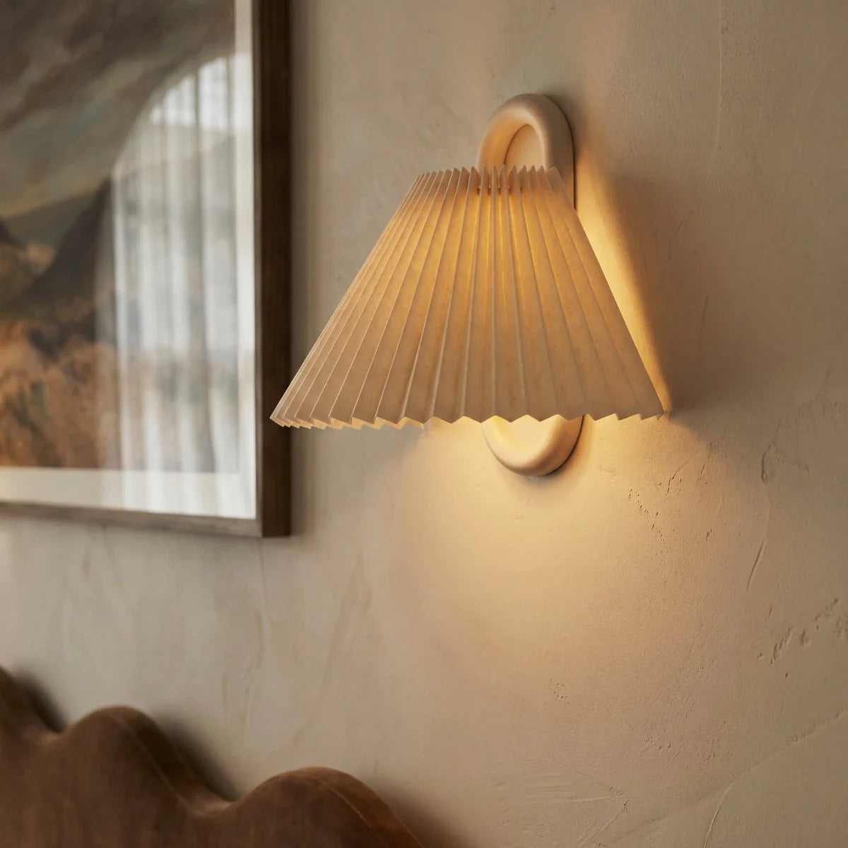 Close-up of a Clérachic pleated cream fabric shade and wood ring bracket, glowing beside a framed painting on the wall.
