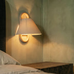 Close-up of a Clérachic wall light with a plain fabric shade, glowing warmly above a bedside table in a green-toned room.