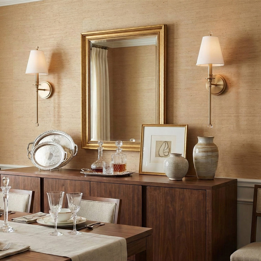 A pair of gold-toned Clérachic sconces flanking a gilt-framed mirror above a walnut sideboard in a dining room with grasscloth walls.