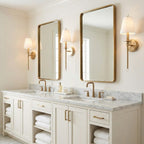 Gold-toned Clérachic sconces beside two brass-framed mirrors above a marble double vanity, with white fabric shades glowing softly.