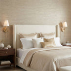 Lit Clérachic wall lights with white shades on both sides of an upholstered cream headboard, over a bed dressed in champagne linen.
