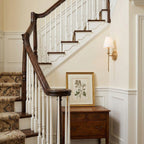 A single gold-toned Clérachic wall light on a stairwell wall, lit above a wooden chest and a framed botanical print.