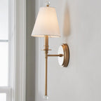 Close view of the tapered white shade, clear glass finial and gold-toned round backplate of this Clérachic wall light on a pale wall.