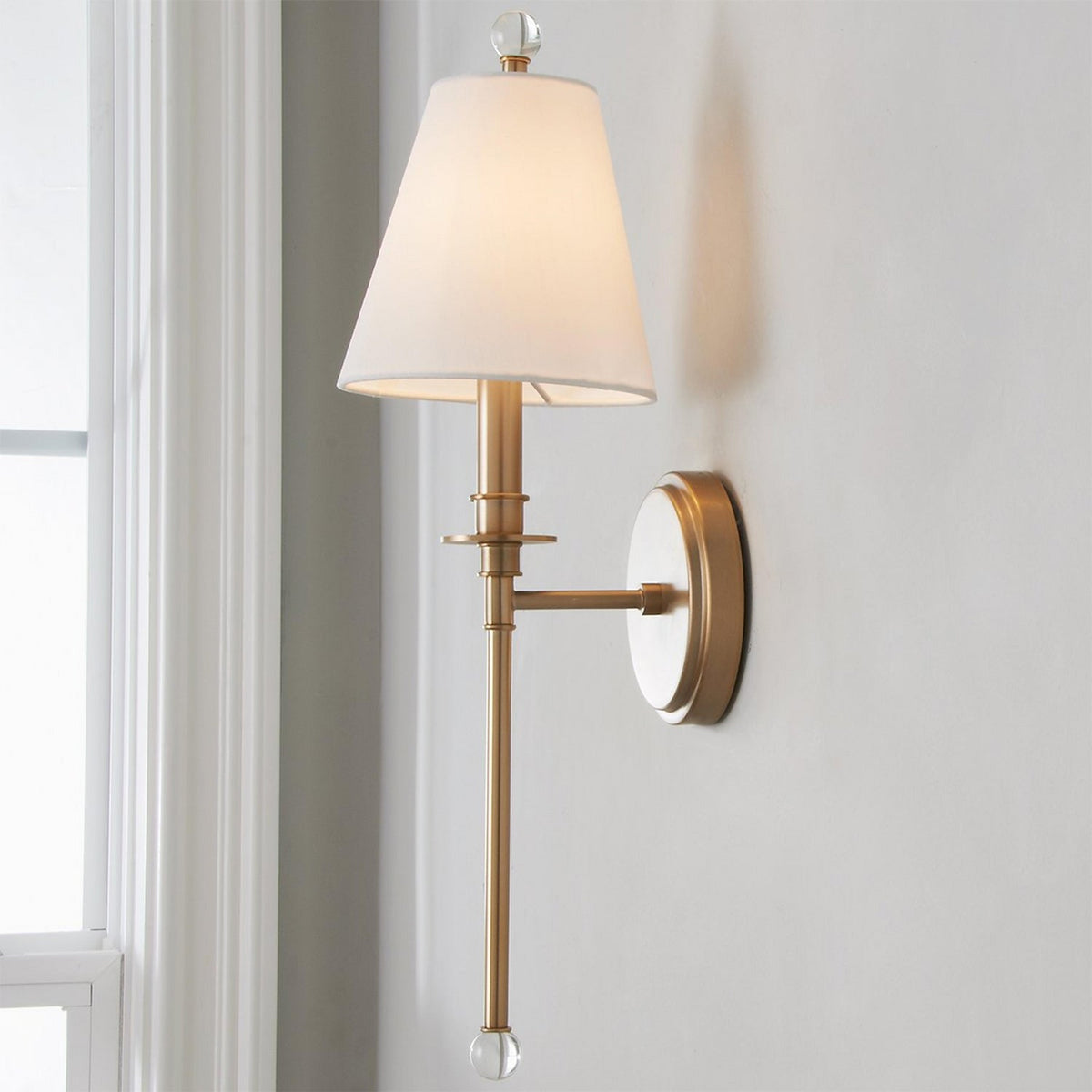 Close view of the tapered white shade, clear glass finial and gold-toned round backplate of this Clérachic wall light on a pale wall.