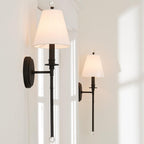Two black Clérachic sconces with white shades at different heights on a bright wall, daylight falling through a window beside them.