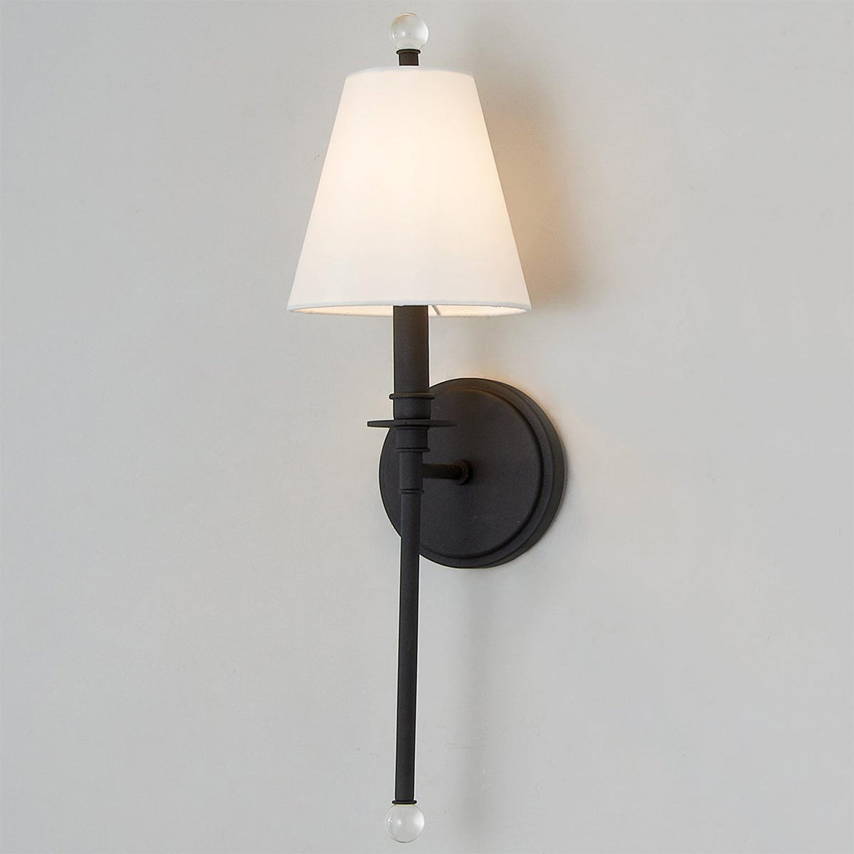 Black Clérachic wall light with a lit white shade and a glass ball at the foot of its slim rod, against a pale grey wall.