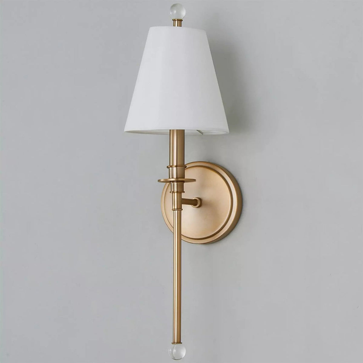 Gold-toned Clérachic wall light with an unlit white fabric shade and a round backplate, mounted on a soft grey wall.