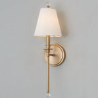 The same gold-toned Clérachic wall light switched on, its white shade glowing warmly against a grey wall.
