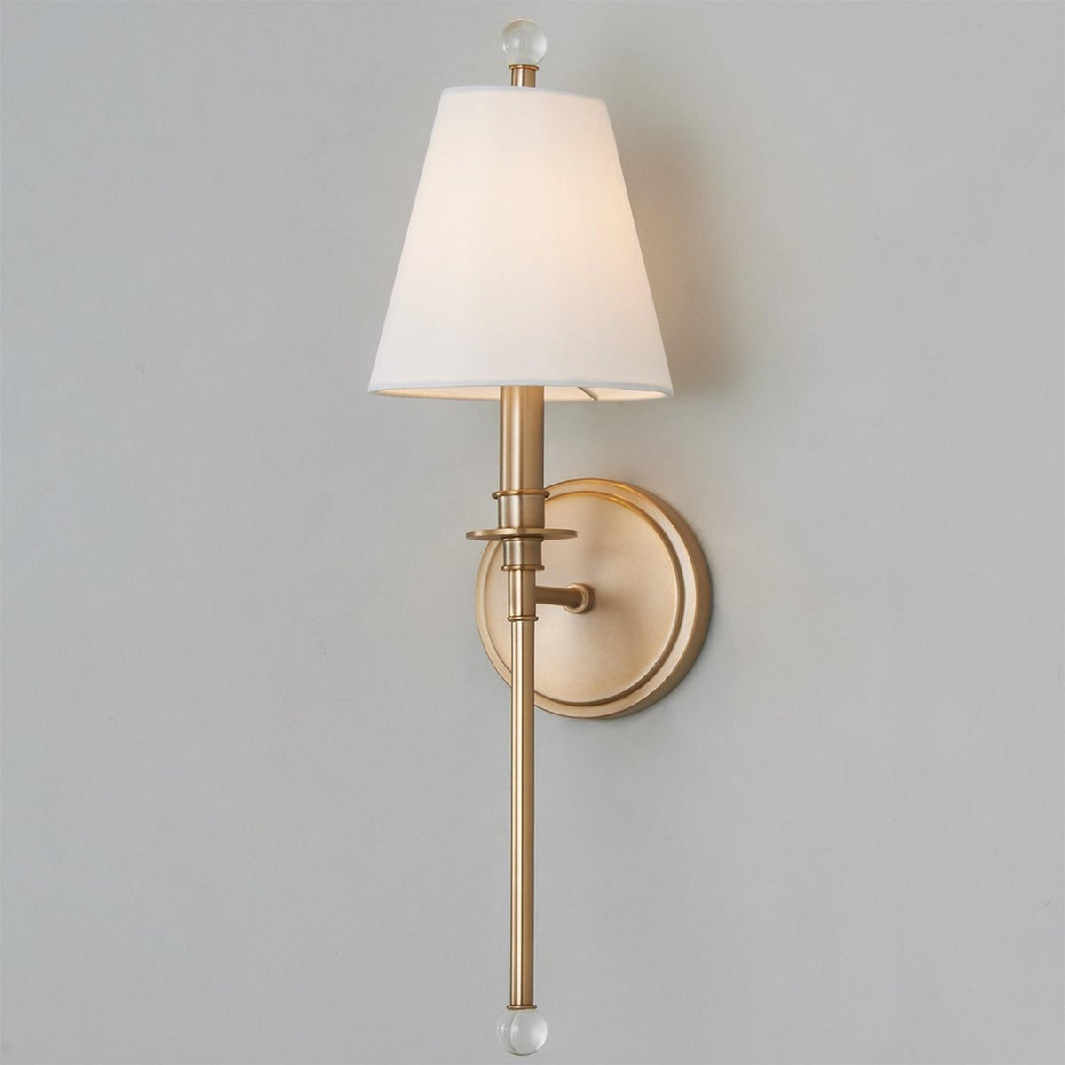 The same gold-toned Clérachic wall light switched on, its white shade glowing warmly against a grey wall.