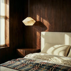 Clérachic alabaster pendant glowing amber beside a bed with a cream headboard and a walnut-panelled wall.