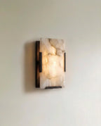 Clérachic single alabaster wall light in dark metal, lit, the veined stone glowing warmly against a pale wall