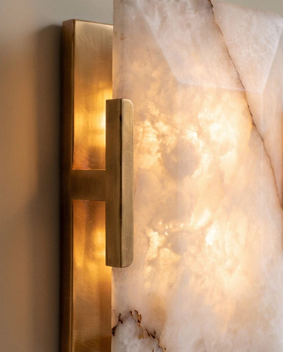 Detail of the Clérachic alabaster wall light's brushed brass T-bracket beside the softly lit, translucent stone panel