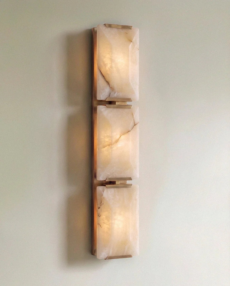 Clérachic three-panel alabaster wall light in brass, lit, the stacked veined panels glowing warm on a sage wall