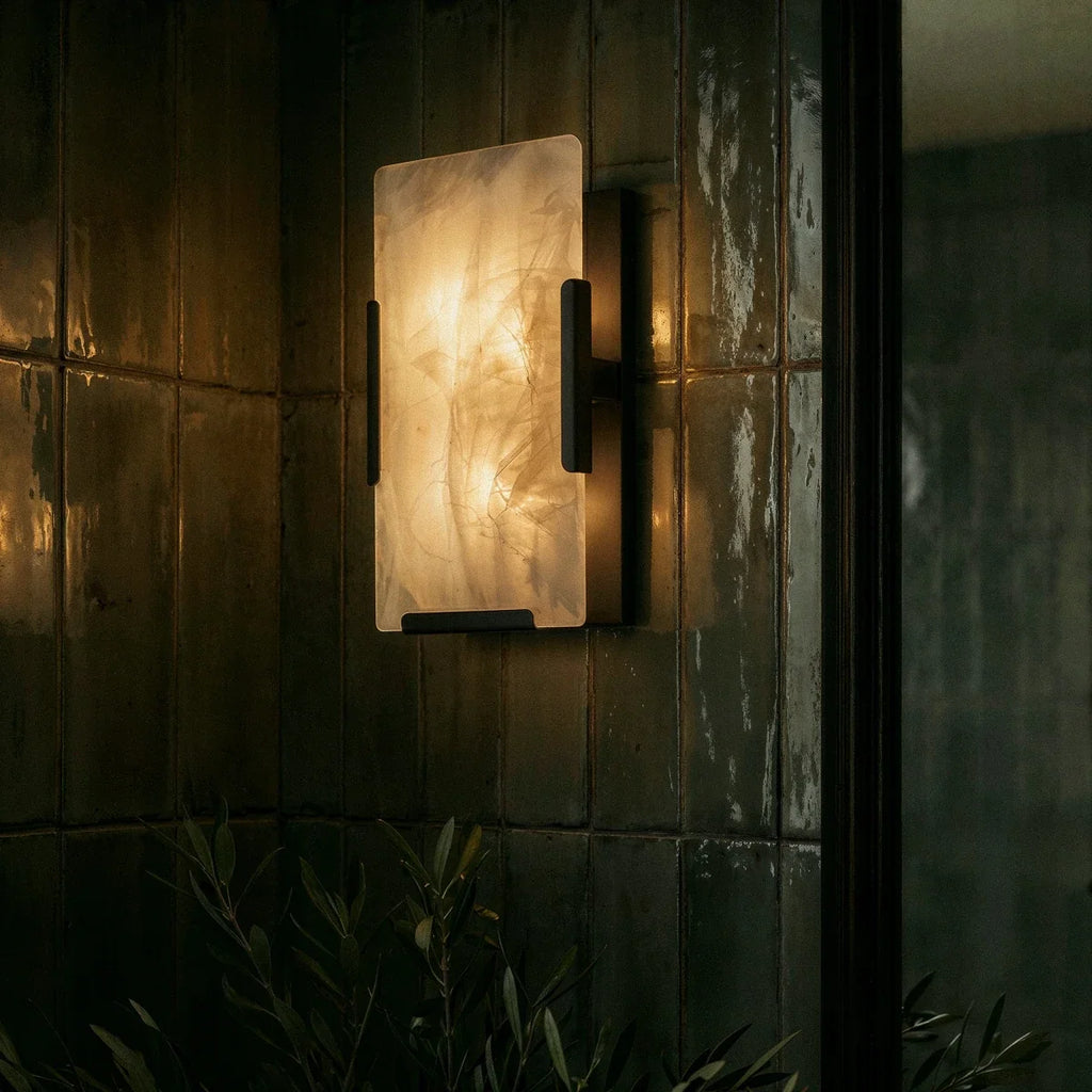 Clérachic alabaster wall light in dark metal glowing on glossy green tiles, with olive leaves in the foreground