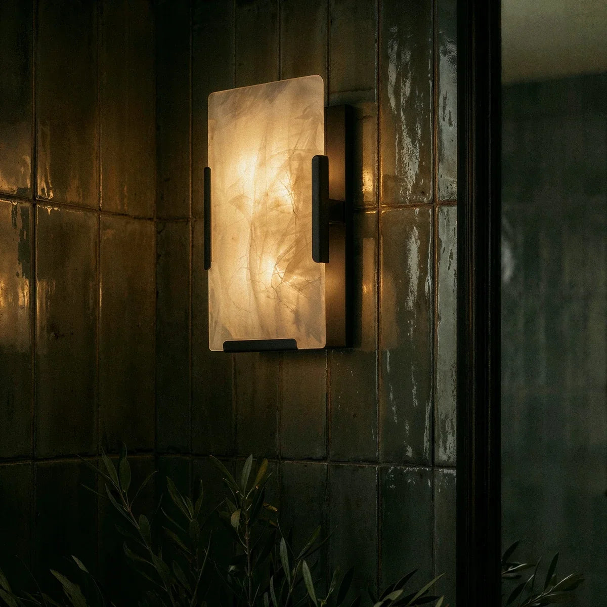 Clérachic alabaster wall light in dark metal glowing on glossy green tiles, with olive leaves in the foreground