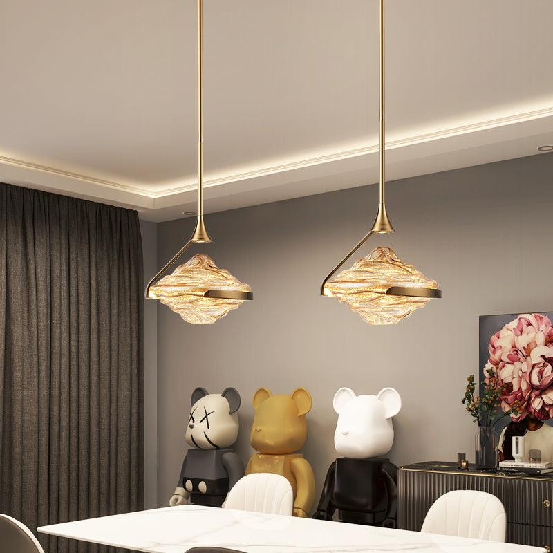 Two faceted amber glass pendants on slim gold rods hang under a lit ceiling edge above a white dining table, from Clérachic.