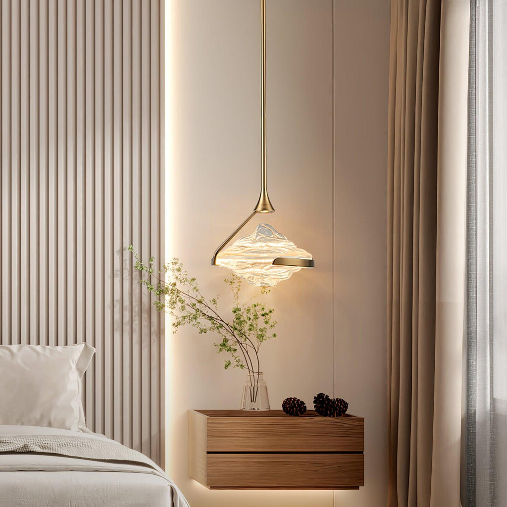 A faceted amber glass pendant on a slim gold rod glows beside a bed, against a timber-slat wall with a floating bedside cabinet, from Clérachic.
