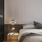 A faceted glass pendant with a chrome rod hangs beside a bed with grey bedding and a small wooden bedside table, from Clérachic.