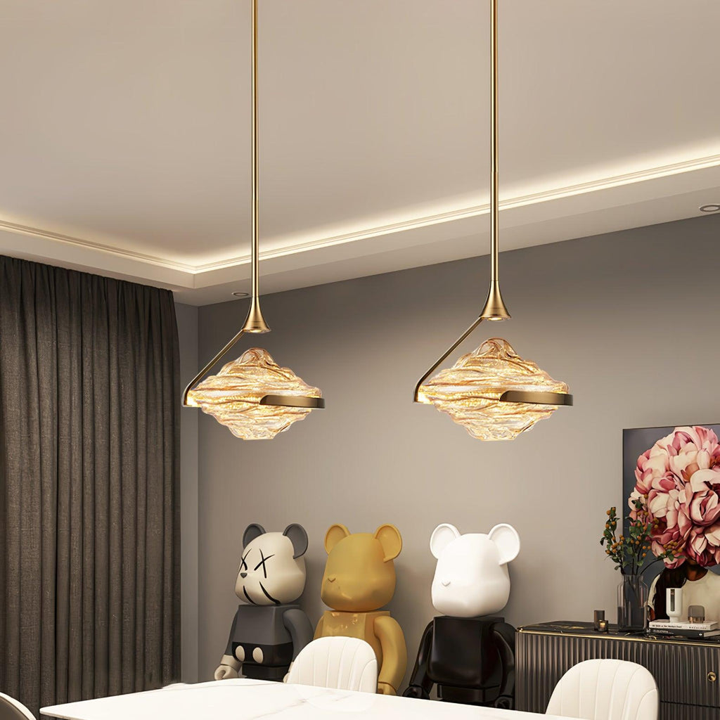 Two faceted amber glass pendants on gold rods hang beneath a lit cove ceiling above a white table and bright chairs, from Clérachic.
