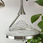 Close view of a rippled clear glass shade with a silver ring and arm, a green leaf at the edge, on this Clérachic pendant.