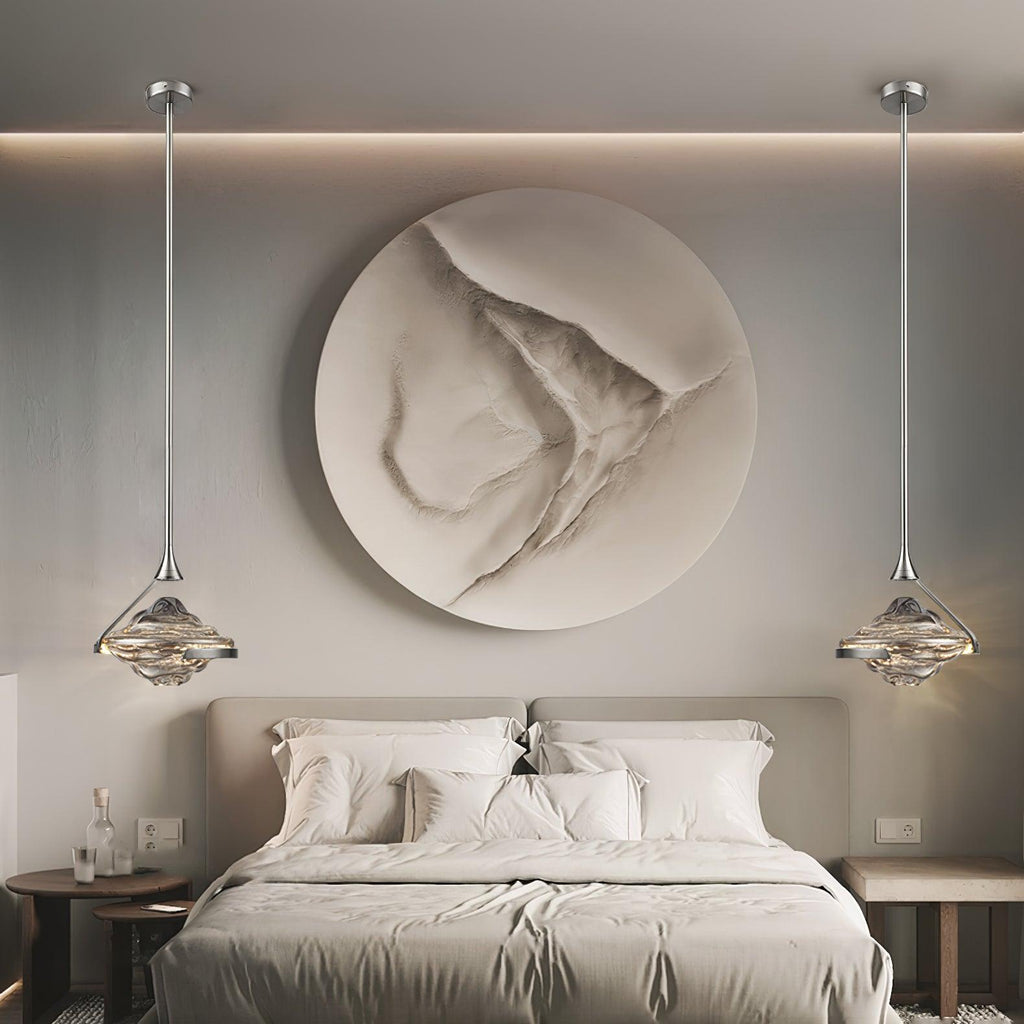 Two faceted glass pendants on slim silver rods hang either side of a bed, in front of a round textured wall panel, from Clérachic.